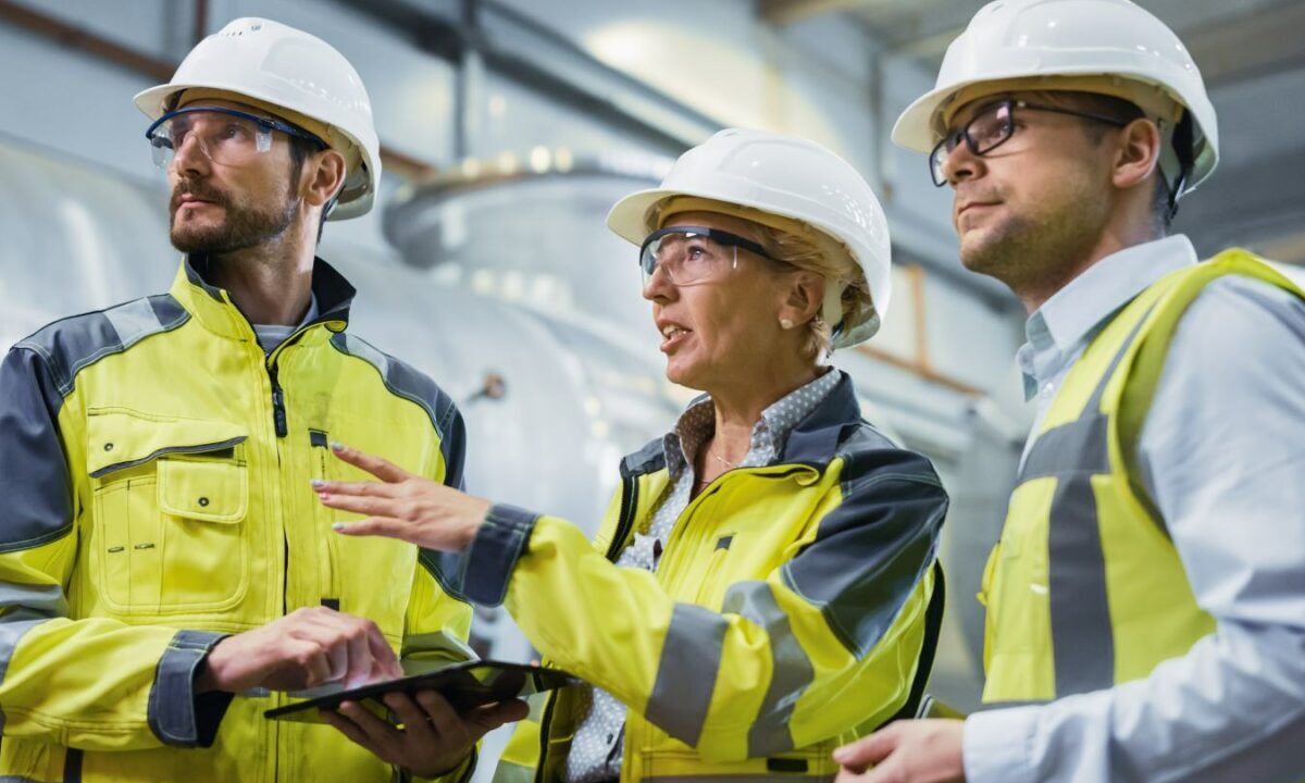 Three Heavy Industry Engineers Stand in Pipe Manufacturing Factory, Use Digital Tablet Computer, Have Discussion. Large Pipe Assembled. Design and Construction of Oil, Gas and Fuels Transport Pipeline