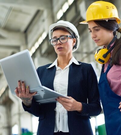 Pensive female building contractor demonstrating online sketch on portable computer to colleague in workwear in factory workshop