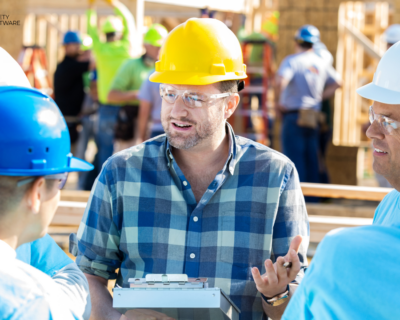 Construction site supervisor wearing a yellow hard hat and safety glasses discusses workplace safety procedures with a group of workers
