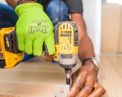A worker wearing a bright green safety glove uses a power drill to fasten a metal bracket to a wooden board, symbolizing how everyday workers quietly reshape safety practices through hands-on choices and small acts of responsibility.
