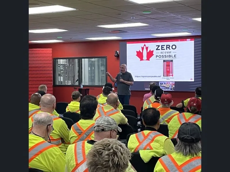 Spencer Beach delivers a workplace safety talk to a room full of workers in high-visibility clothing.