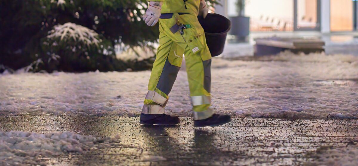 Worker in reflective PPE spreading grit on frozen pavement as part of winter walkway safety.
