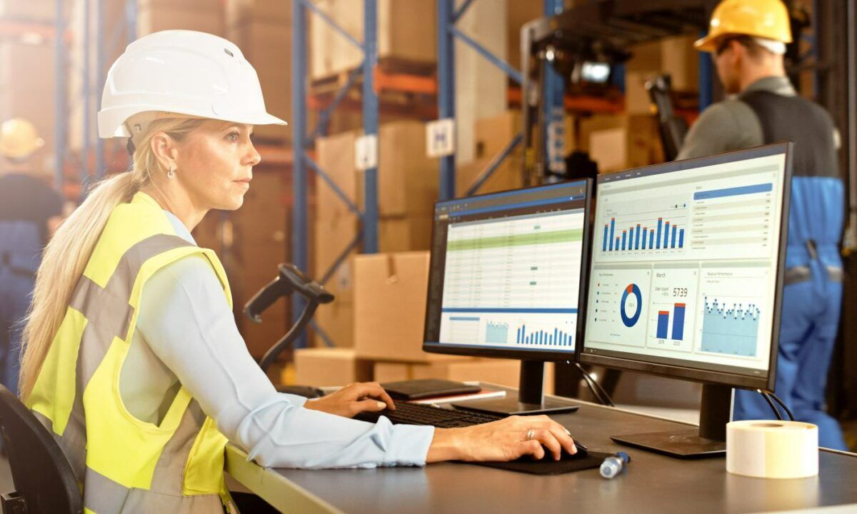 Professional Female Worker Wearing Hard Hat Uses Computer with Inventory Status Checking and Delivery Software in the Retail Warehouse full of Shelves with Goods. Delivery, Distribution Center