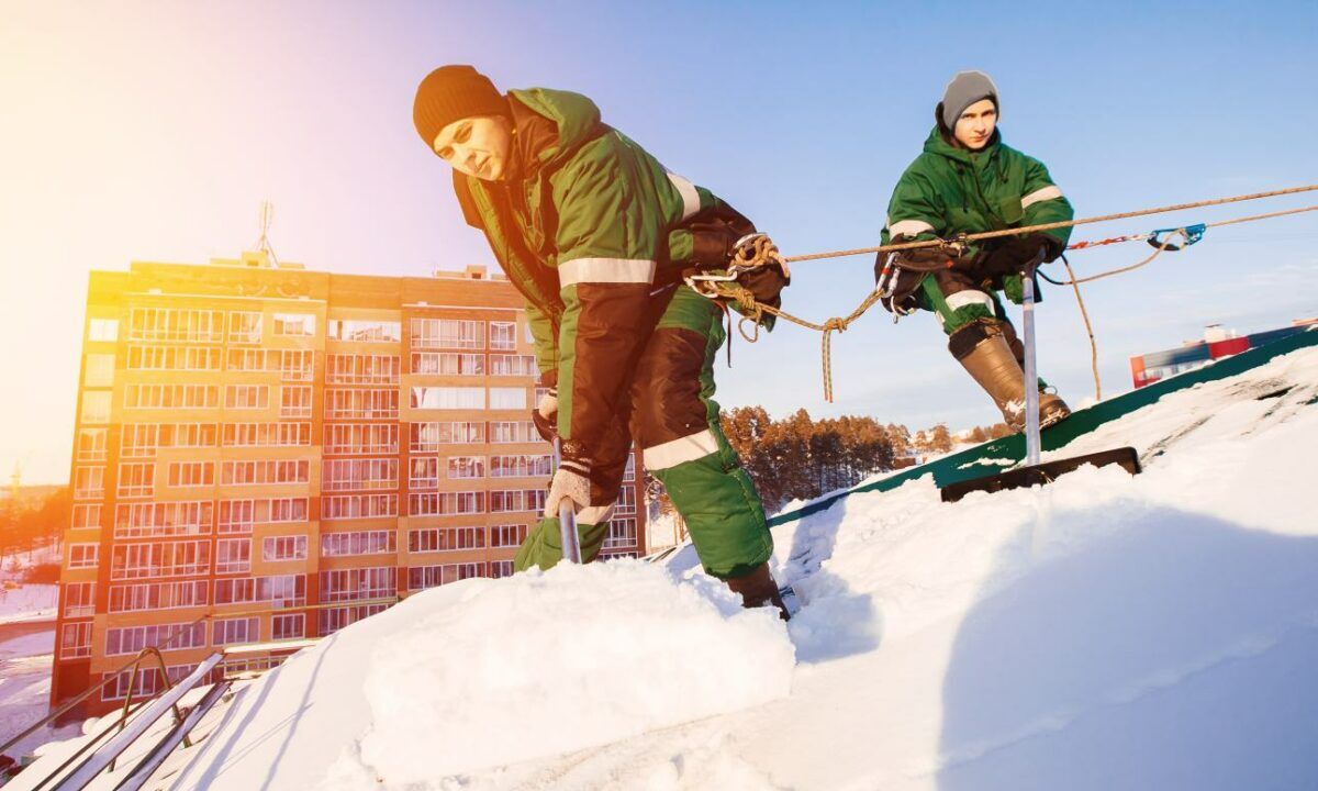 Snow cleaning. Team of male workers clean roof of building from snow with shovels in securing belts of mantra.