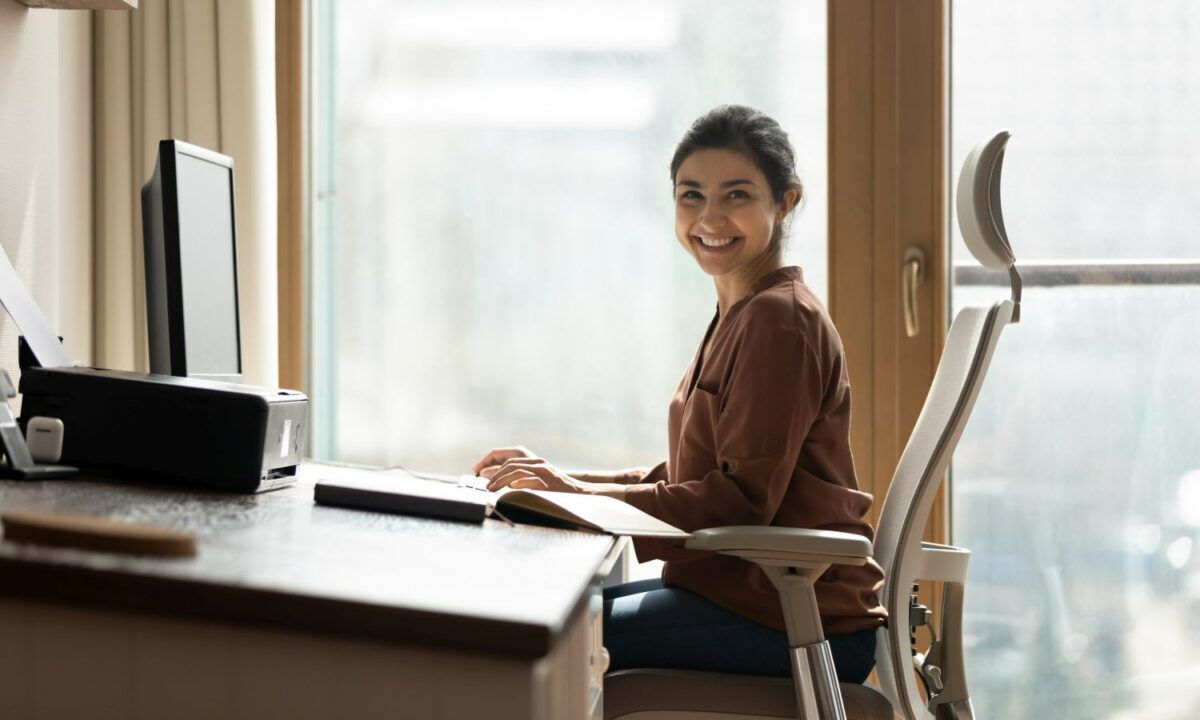 Modern day worker. Portrait of happy biracial business woman freelancer sit by computer at comfy workplace at corporate workspace or at home. Smiling young indian lady office employee look at camera