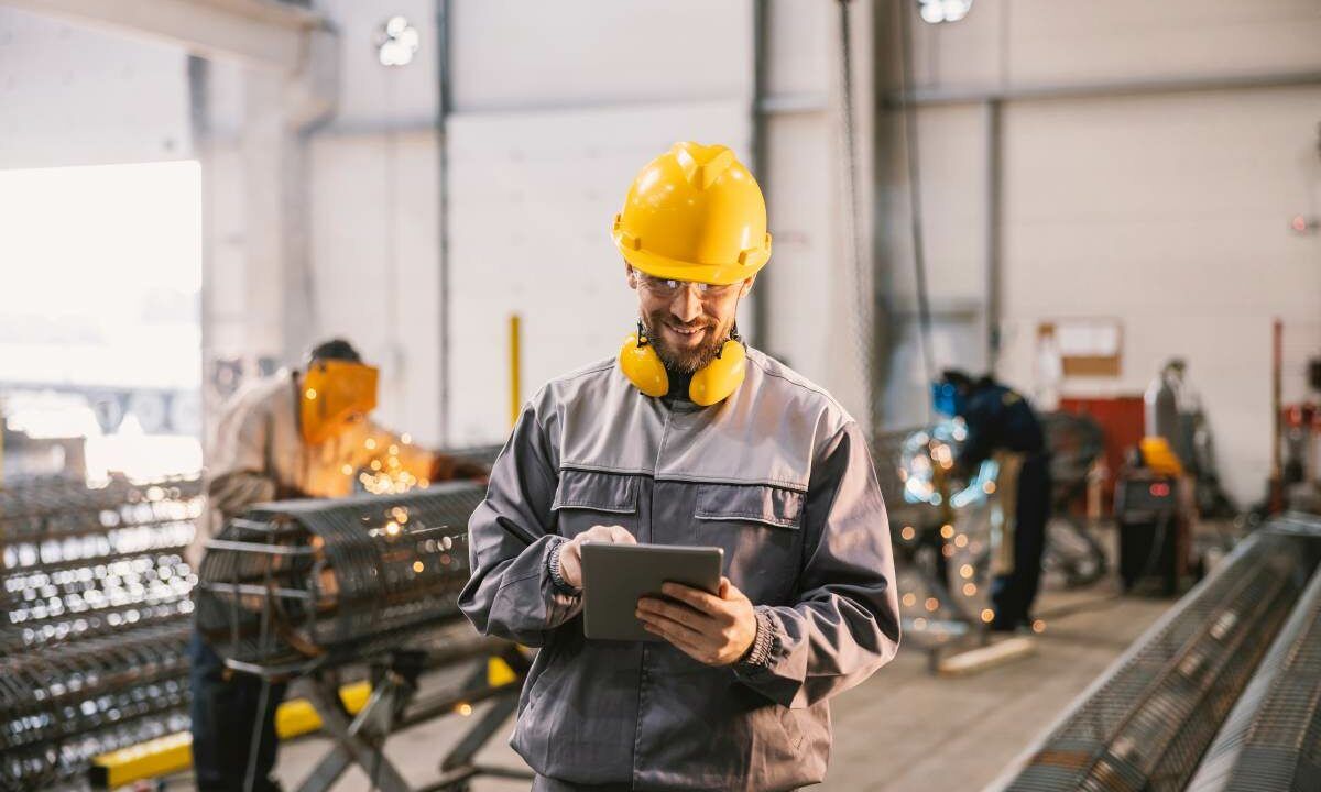 A heavy industry supervisor is taking course on tablet in facility while smiling at the camera. In blurry background workers welding and grinding metal framework.