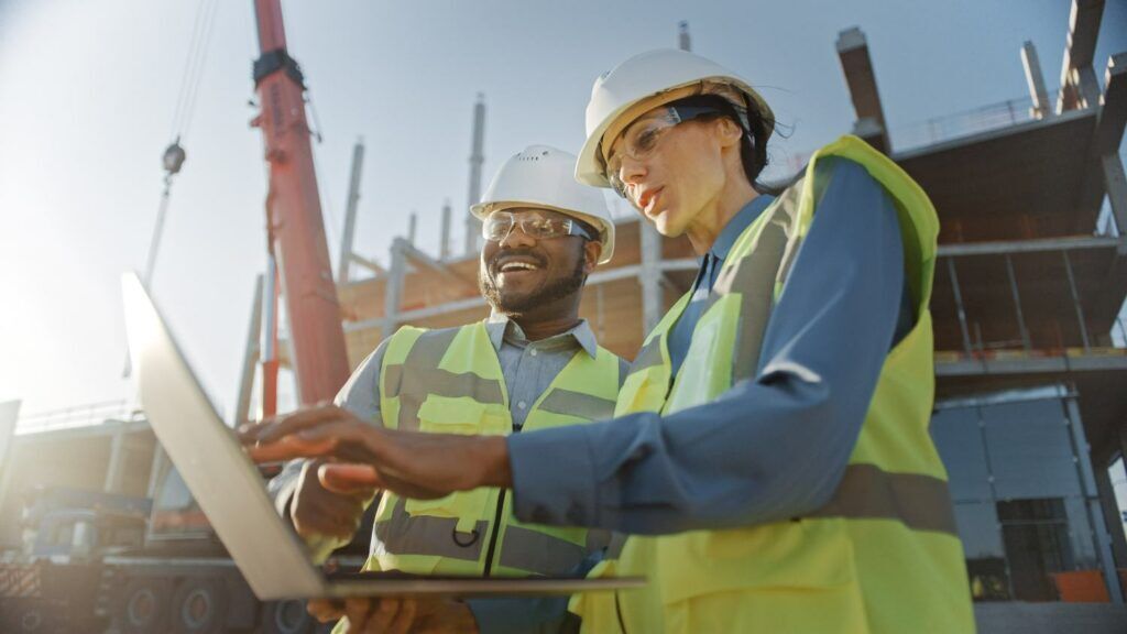 Two Specialists Inspect Commercial, Industrial Building Construction Site. Real Estate Project with Civil Engineer, Investor Use Laptop. In the Background Crane, Skyscraper Concrete Formwork Frames