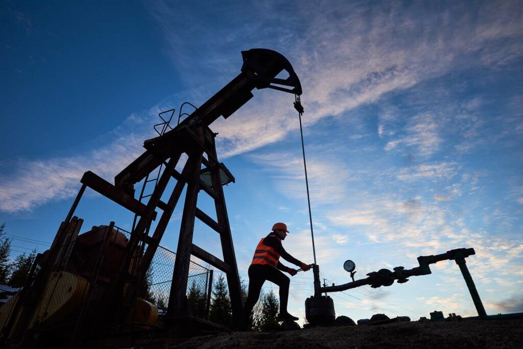 Silhouette of engineer in work vest and helmet working on petroleum pump jack. Oil worker using oil pump rocker-machine in oil field under evening sky. Concept of oil extraction and petroleum industry