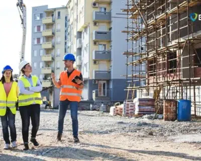 Construction worker in a high-visibility vest and safety harness stands between concrete columns, looking toward the sunset, symbolizing hope and progress in a developing site.